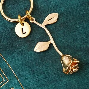 May include: Gold rose keychain with a letter L charm. The rose is detailed with a stem and leaves. The keychain is attached to a gold key ring.