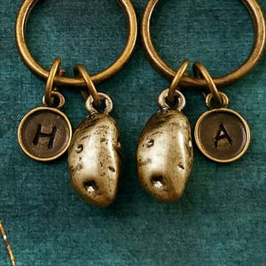 May include: Two gold-toned keychains with potato charms and letter charms. The charms are attached to key rings. The letter charms are engraved with the letters "H" and "A".