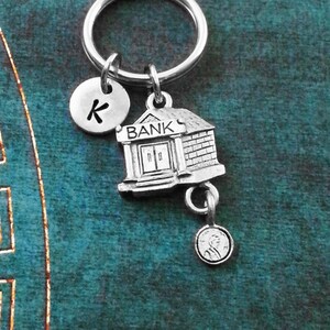 May include: Silver-toned keychain featuring a miniature bank building charm, a round charm with the letter "K", and a small coin charm. The bank charm has the word "BANK" above the doors. The keychain is attached to a silver key ring.