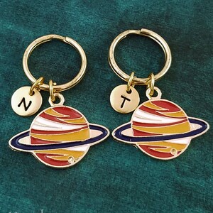 May include: Two gold keychains with a red, white, and yellow planet charm with a blue ring around it. The charms are personalized with the letters "N" and "T".