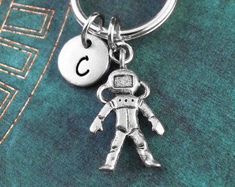 May include: Silver astronaut keychain with a silver initial charm with the letter C. The keychain is on a teal background.