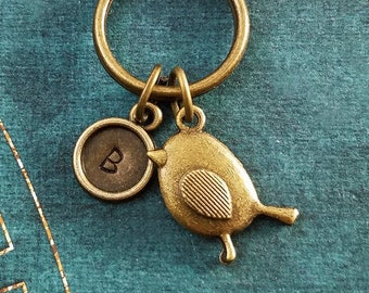May include: A gold-toned keychain with a small bird charm and a round charm with the letter "B" engraved on it. The keychain is on a teal background.
