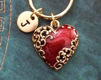 May include: Gold heart-shaped keychain with a red enamel finish and intricate gold filigree details. A small gold disc with the letter 'J' is attached to the keychain.