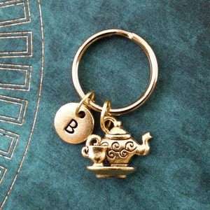 May include: Gold-colored keychain featuring a teapot and teacup charm, a round charm with the letter "B", and a keyring. The charms are detailed with decorative swirls. The keychain is on a teal background.