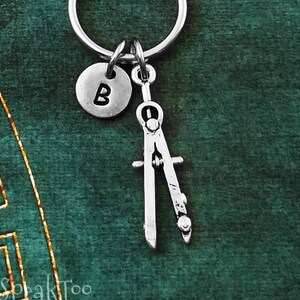 May include: Silver compass keychain with a silver initial charm. The initial charm has the letter "B" on it. The keychain is on a green background.