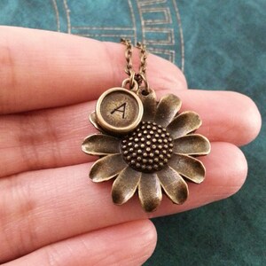 May include: A bronze necklace with a sunflower charm and a typewriter key charm with the letter 'A' on it.