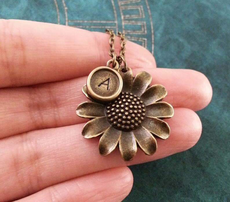 May include: A bronze necklace with a sunflower charm and a typewriter key charm with the letter 'A' on it.