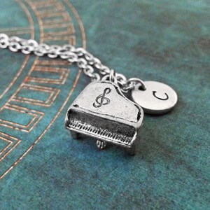 Op de afbeelding: Een zilveren ketting met een miniatuur pianobedel en een zilveren bedel met de letter "C" erop gegraveerd.