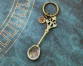 May include: A gold-toned spoon keychain with a decorative floral design and a small round charm with the letter 'A' on it. The keychain is attached to a silver key ring.