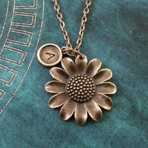 May include: A bronze necklace with a sunflower charm and a small initial charm. The initial charm is engraved with the letter "A".