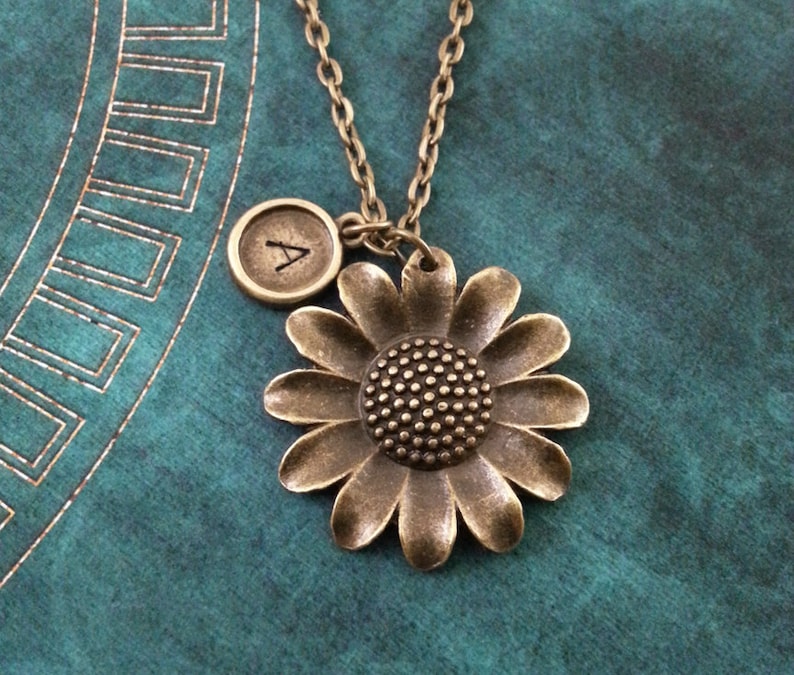May include: A bronze necklace with a sunflower charm and a small initial charm. The initial charm is engraved with the letter "A".