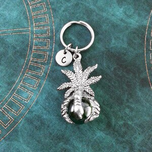 May include: Silver keychain featuring a marijuana leaf design with a green glass bead. The keychain has a silver letter 'C' charm.