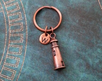 May include: A copper-colored keychain featuring a lighthouse charm and a round charm with the letter "W". The keychain is attached to a copper-colored ring. The background is a teal-colored surface.