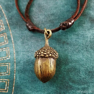 May include: A bronze-colored acorn pendant necklace with a brown leather cord. The acorn has a detailed cap and a textured surface. The necklace is displayed against a teal background.