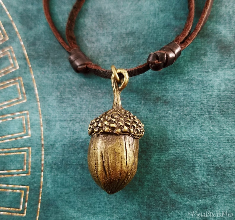 May include: A bronze-colored acorn pendant necklace with a brown leather cord. The acorn has a detailed cap and a textured surface. The necklace is displayed against a teal background.