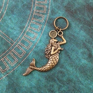 May include: A gold-toned mermaid keychain with a small round charm with the letter "E" on it. The mermaid is holding the charm in her hand. The keychain is on a teal background.