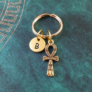 May include: Gold-toned keychain with a small round charm with the letter "B" and a larger charm shaped like an ankh, an ancient Egyptian symbol.