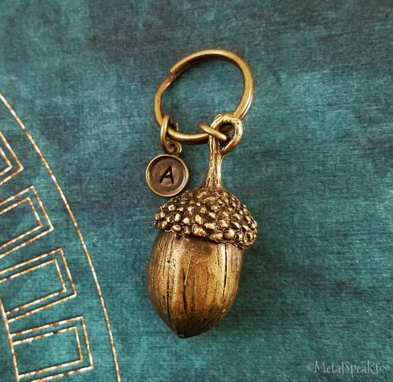 May include: A bronze-colored acorn keychain with a round charm bearing the letter "A". The acorn has a textured cap and a smooth body, attached to a keyring. The background is a teal-colored fabric.