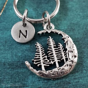 May include: Silver-toned keychain featuring a crescent moon design with three detailed pine trees. A small, round charm with the letter "N" is also attached. The keychain is designed for keys and accessories.