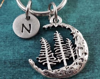 May include: Silver-toned keychain featuring a crescent moon design with three detailed pine trees. A small, round charm with the letter "N" is also attached. The keychain is designed for keys and accessories.