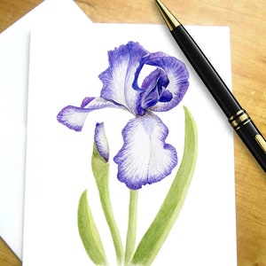 May include: A watercolor painting of a purple and white iris flower with green leaves and a bud. The flower is on a white card with a white envelope, and a black pen rests on a wooden surface.