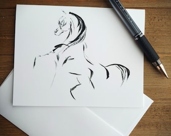 May include: A black and white ink drawing of a horse on a white card. The horse is depicted with flowing lines, suggesting movement. A black pen with a silver barrel rests on the card. The card is on top of a white envelope.