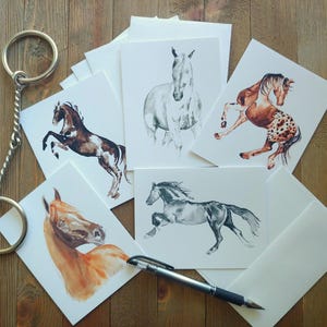 May include: A collection of watercolor horse art prints and blank cards. The prints feature various horse poses and colors, including brown, black, and white. A silver key ring and pen are also present. The cards are arranged on a wooden surface.