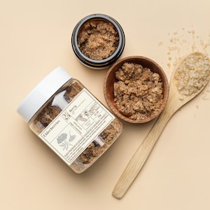 Può includere: Un barattolo di vetro pieno di scrub allo zucchero di canna con un'etichetta che recita "Elderberries Berry Sugar Scrub 125g $11.50". Il barattolo è su una superficie marrone chiaro accanto a una piccola ciotola di legno piena di scrub allo zucchero di canna e un cucchiaio di legno con cristalli di zucchero bianco.