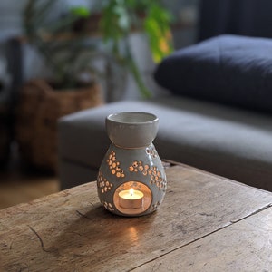 May include: A grey ceramic essential oil diffuser with a lit tea light candle inside. The diffuser has a bulbous base with decorative cut-out patterns and a smaller top section. It sits on a rustic wooden table, with a blurred background of a sofa and plants.
