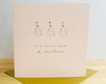 Puede incluir: Tarjeta navideña rosa pálido con tres árboles de Navidad dorados y el texto "to a lovely mum at christmas". La tarjeta está sobre un sobre dorado.