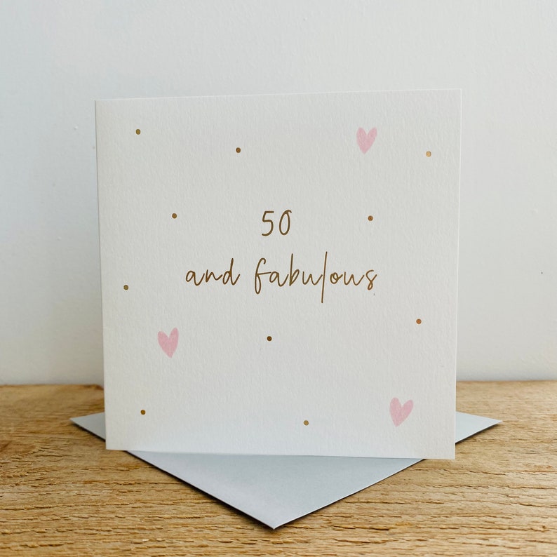 May include: A white card with gold lettering that says "50 and fabulous". The card is decorated with small gold dots and pink hearts.