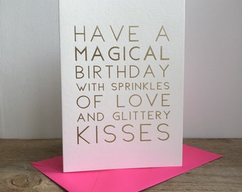 Puede incluir: Una tarjeta blanca con letras doradas que dice "Have a magical birthday with sprinkles of love and glittery kisses". La tarjeta está sobre un sobre rosa.