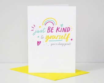 May include: A white greeting card with the text "just BE KIND to yourself, you're doing great!" in colourful lettering. The card features a rainbow, hearts, and stars. It rests on a yellow envelope.