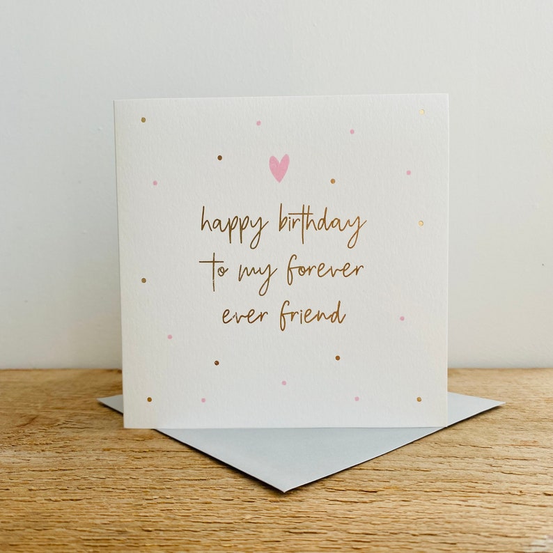Puede incluir: Una tarjeta blanca con letras doradas que dice "happy birthday to my forever ever friend". La tarjeta tiene un coraz&oacute;n rosa y puntos rosas y dorados.
