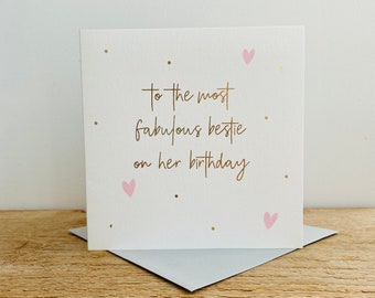 May include: A white card with gold lettering that reads "To the most fabulous bestie on her birthday". The card has pink hearts and gold dots scattered across the front.