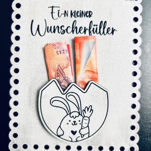 May include: A white wooden card with a scalloped edge. The card has a black and white illustration of a bunny holding a carrot. The text "Ein kleiner Wunscherschfiller" is written in black on the card. There are two folded bills in the pocket of the card.