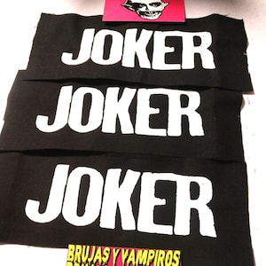 Pode incluir: Três remendos de tecido preto com a palavra "JOKER" impressa em letras brancas. Os remendos estão empilhados um em cima do outro. Um cartão rosa com um monstro de desenho animado é visível no fundo. O cartão também tem o texto "BRUJAS Y VAMPIRIOS" impresso nele.