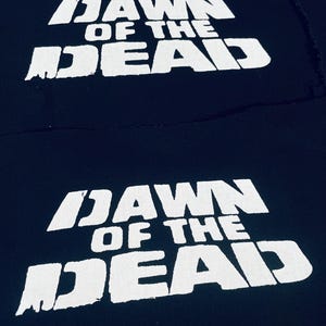 Könnte beinhalten: Zwei schwarze Stoff-Patches mit weißem Text, der "Dawn of the Dead" lautet.