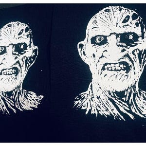 May include: Black fabric featuring a white graphic of a grotesque face with a wide, toothy grin. The image shows two identical prints of the face, side by side. The face has a scarred appearance and a menacing expression.