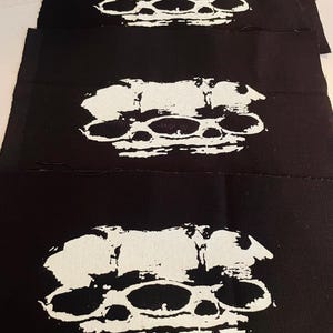 May include: Three black fabric patches, each featuring a white brass knuckles design. The graphic design is a bold representation of the weapon. The patches are rectangular and made of a sturdy material, suitable for sewing or ironing.