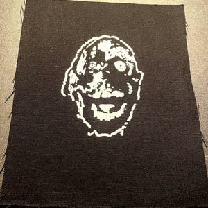 May include: A black fabric patch with a white, distorted face illustration. The face has a wide grin and a single visible eye. The patch is rectangular with frayed edges.