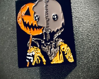 Op de afbeelding: Zwarte en oranje emaille pin met een personage met een gestikt gezicht dat een jack-o'-lantern vasthoudt. Het personage heeft twee zwarte knopen als ogen.