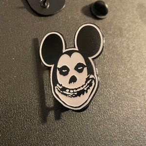 以下が含まれることがあります： ミッキーマウスの耳が付いた頭蓋骨が描かれた黒と白のエナメルピン。頭蓋骨は歯を見せて笑っています。