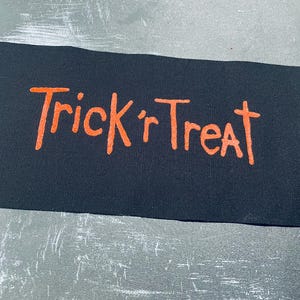 May include: A rectangular black fabric patch featuring the text Trick 'r Treat printed in a bright orange, hand-drawn style font. The patch has a visible woven texture and clean, straight edges, presenting a bold contrast between the dark background and the vibrant orange lettering.