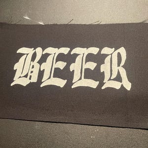 Op de afbeelding: Een zwarte stoffen patch met het woord "BEER" in wit gedrukt in een gotisch lettertype.