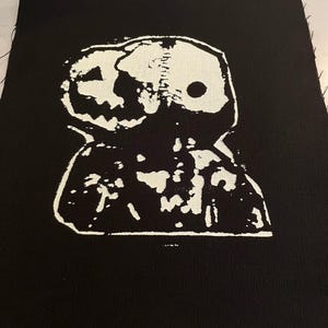 May include: A black fabric patch with a white design of a cartoon character with a skull-like head and a large, toothy grin.
