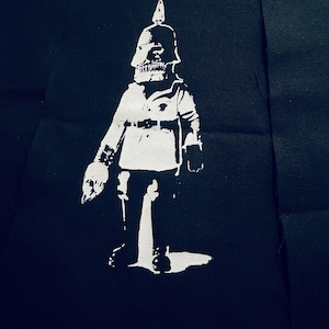 Op de afbeelding: Een wit silhouet van een figuur die een helm en uniform draagt, die lijkt op een soldaat of een bewaker, op een zwarte stoffen achtergrond.