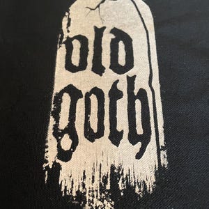 Op de afbeelding: Een zwarte stoffen patch met een witte, verweerde, grafsteen-vormige afbeelding met de tekst "old goth".