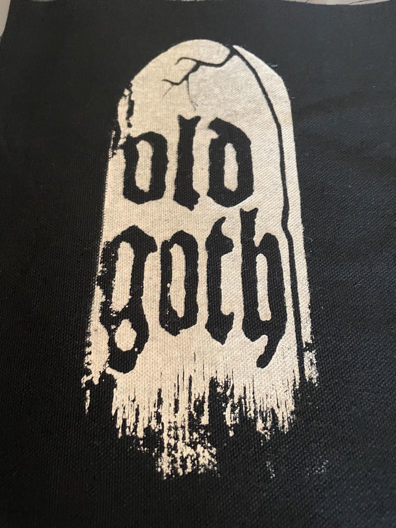 Op de afbeelding: Een zwarte stoffen patch met een witte, verweerde, grafsteen-vormige afbeelding met de tekst "old goth".
