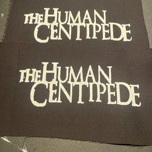 Könnte beinhalten: Zwei schwarze Stoffpatches mit weiß gesticktem Text. Der Text lautet "The Human Centipede".